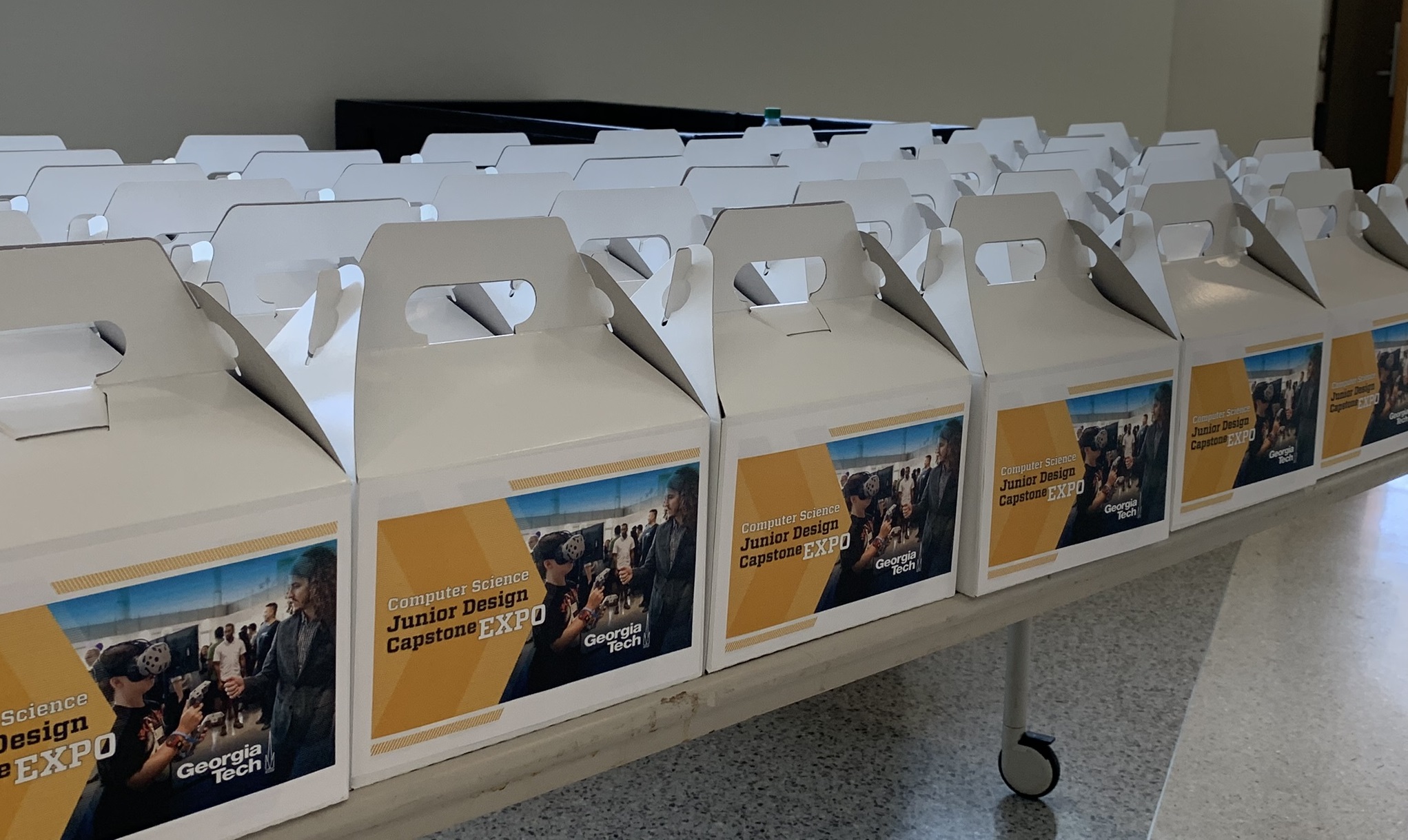 Rows of lunchboxes are lined up on a table. Each box has a label with the words "Junior Design Capstone Expo" and an image of two people trying virtual reality technology.