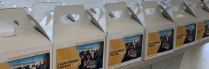 Rows of lunchboxes are lined up on a table. Each box has a label with the words "Junior Design Capstone Expo" and an image of two people trying virtual reality technology.