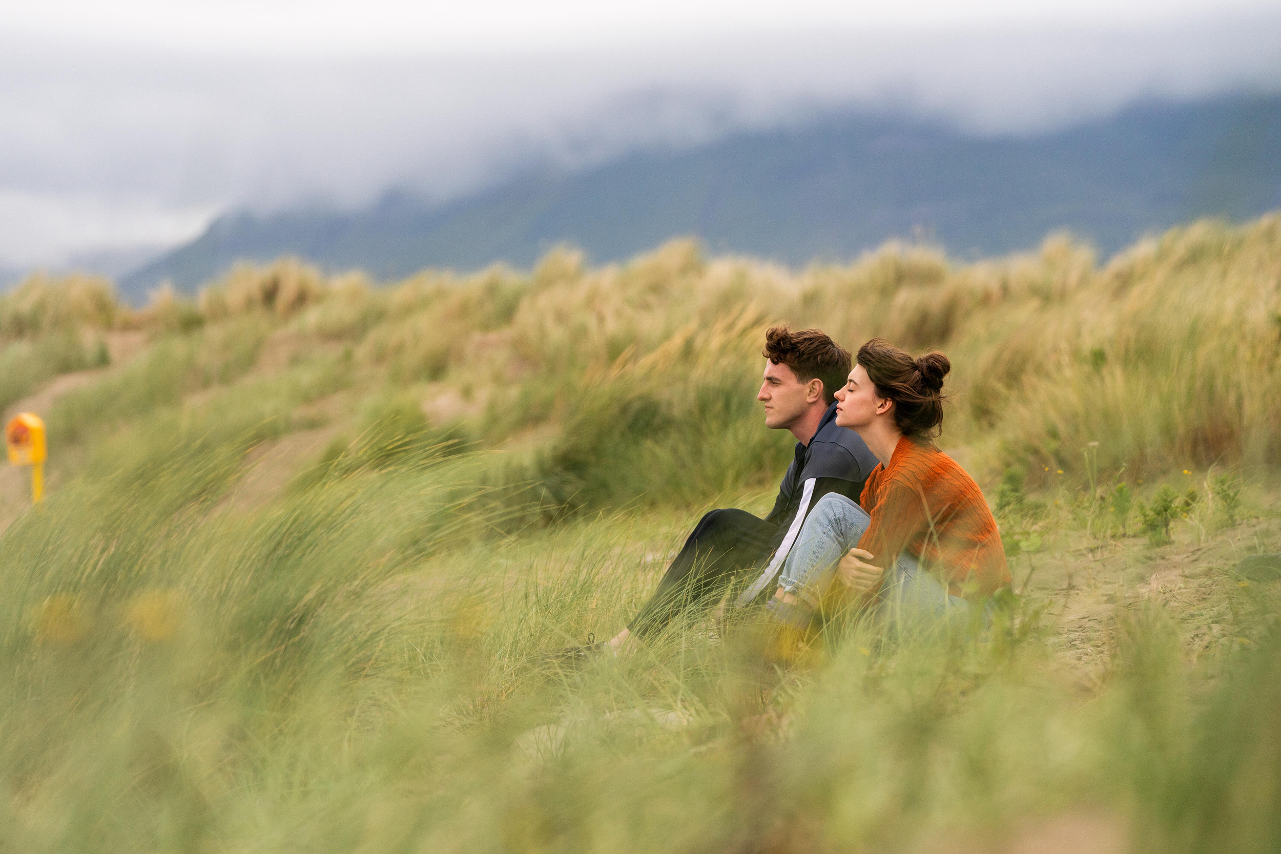 Actress Daisey Edgar-Jones (as Marianne) and actor Paul Mescal (as Connell) sit together in tall, green grass and face an on-coming wind in this still from the Hulu show "Normal People."