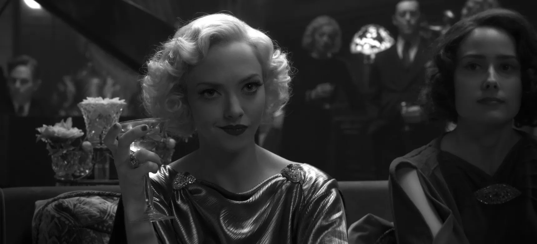 Amanda Seyfried, as Marion Davies in Mank, sits at a party in formal dress. Sheis holding a martini and has a wry smile on her face.